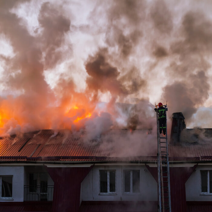 Firefighters on a ladder extinguishing a blazing fire on the roof of an apartment building in the city, with smoke billowing from the flames.