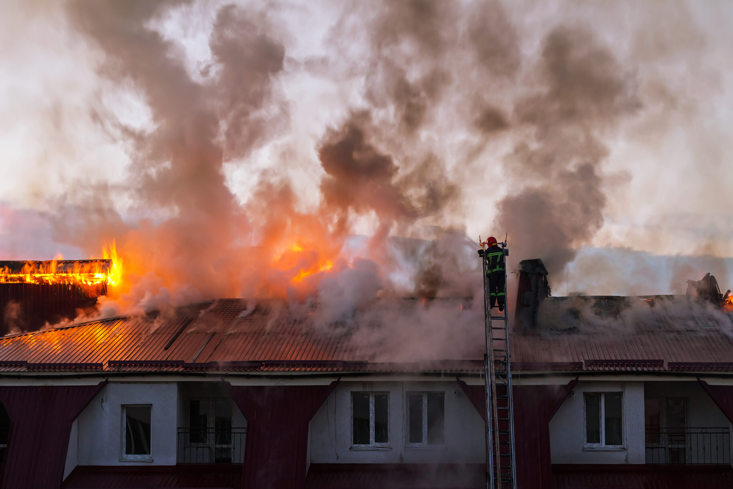 Firefighters on a ladder extinguishing a blazing fire on the roof of an apartment building in the city, with smoke billowing from the flames.